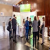 Das Bild zeigt eine Gruppe von Personen, die sich vor einem Ausstellungsstand auf einer Konferenz oder Messe unterhalten. 