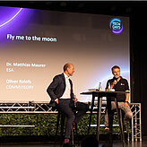 Das Bild zeigt zwei Männer die auf einer Bühne etwas vortragen