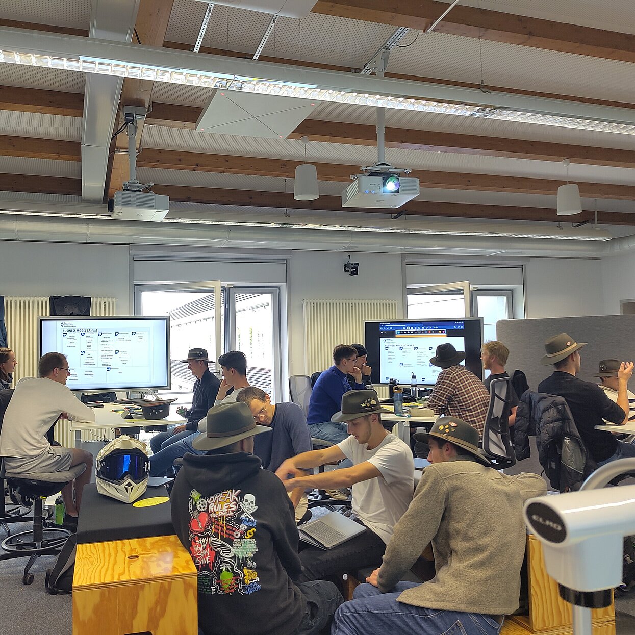 Studierende arbeiten in 4-5 Gruppen und sitzen an Tischen und Monitoren. Einige tragen gründe Hüte. 