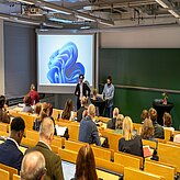 Vortragssituation in einem Hörsaal mit mehreren sitzenden Zuhörern und zwei stehenden Personen vor einer Leinwand und einer grünen Tafel