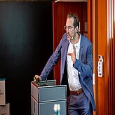 Eine Person im Anzug steht hinter einem Rednerpult und hält möglicherweise eine Präsentation oder Rede. Die Person hält ein Mikrofon in der Hand