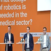 Drei Personen stehen auf einer Bühne hinter Rednerpulten. Der Hintergrund ist orange