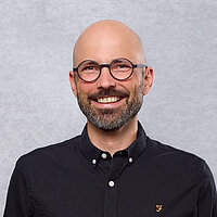 Das Bild zeigt ein Porträt einer lächelnden Person mit Glatze, rundem Brillengestell und kurzem Bart vor einem hellgrauen Hintergrund. Die Person trägt ein dunkles Hemd und blickt direkt in die Kamera. Insgesamt wirkt das Foto freundlich, professionell und klar gestaltet.