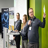 Auf dem Bild sind zwei Frauen und ein Mann zu sehen, welche alle den gleichen Hoodie tragen. Der Mann hält ein Mikrofon und Moderationskarten in der Hand. Eine der Frauen schreibt etwas am Laptop.