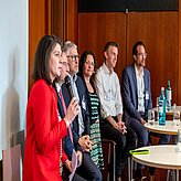 Fünf Personen sitzen auf Stühlen in einem Raum mit Holzverkleidung. Eine Person in roter Jacke hält ein Mikrofon und scheint zu sprechen. Auf dem Tisch vor ihnen stehen Wasserflaschen und Gläser. 