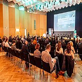Bild zeigt eine Konferenz oder ein Seminar in einem großen Saal. Zahlreiche Personen sitzen in Reihen und blicken auf eine Bühne, auf der eine große Leinwand mit Präsentationsfolien zu sehen ist. 
