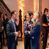 "Mehrere Personen in formeller Kleidung stehen beisammen und unterhalten sich bei einer Veranstaltung. Einige halten Getränke in der Hand. Im Hintergrund ist eine Treppe mit dekorativem Geländer sowie warm beleuchtete Wände zu sehen."