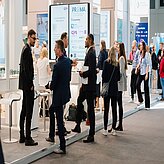 Mehrere Personen stehen und unterhalten sich in einem hellen, modernen Ausstellungsbereich mit Informationsständen