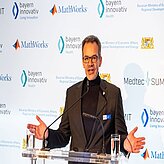 Eine Person steht an einem Rednerpult und hält eine Rede oder Präsentation. Im Hintergrund sind mehrere Logos und Schriftzüge zu sehen,