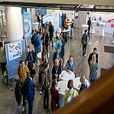 Menschen in einer Halle mit Informationsständen und Stehtischen bei einer Veranstaltung zur Zukunft der Pflege
