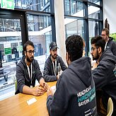 Das Bild zeigt eine Gruppe von fünf Männern, die um einen Holztisch in einem modern wirkenden Raum sitzen. Sie scheinen in ein Gespräch vertieft zu sein. Die Männer tragen dunkelblaue Hoodies mit dem Aufdruck "Healthcare Hackathon Bayern München".
