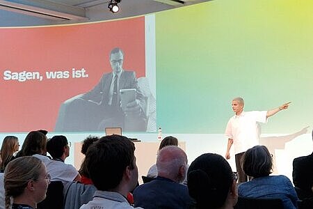 Zu sehen ist der Referent Fabian Grischkat, wie er auf der Bühne steht und mit der linken Hand eine Geste in Richtung seiner Präsentation macht. Er wirkt dabei sehr konzentriert. Die Menschen im Publikum sind nur von hinten zu erkennen, wirken aber interessiert. 