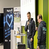 Auf dem Bild sind zwei Herren in grauen Hoodies zu sehen, welche beide ein Mikro in der Hand und einen Vortrag halten.