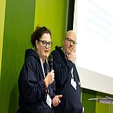 Eine Frau und ein Mann halten einen Vortrag, sie stehen vor grünem Hintergrund