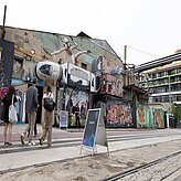 Mehrere Personen gehen an einem ehemaligen Industriegebäude mit großflächigen Graffiti entlang. Im Vordergrund weist ein Aufsteller auf den Check-in der Veranstaltung „TECH DAYS“ hin.
