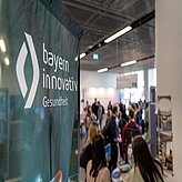 Banner mit dem Schriftzug 'bayern innovativ Gesundheit' vor unscharfem Hintergrund mit mehreren Personen in einem Innenraum