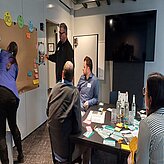 Auf diesem Bild ist eine Gruppe von Menschen in einem Büro oder Besprechungsraum zu sehen. Eine Person steht in der Nähe eines Whiteboards, auf dem verschiedene Notizen und Diagramme zu sehen sind. Eine andere Person sitzt an einem Tisch mit mehreren Laptops und Papieren, und eine weitere Person steht in der Nähe des Tisches.