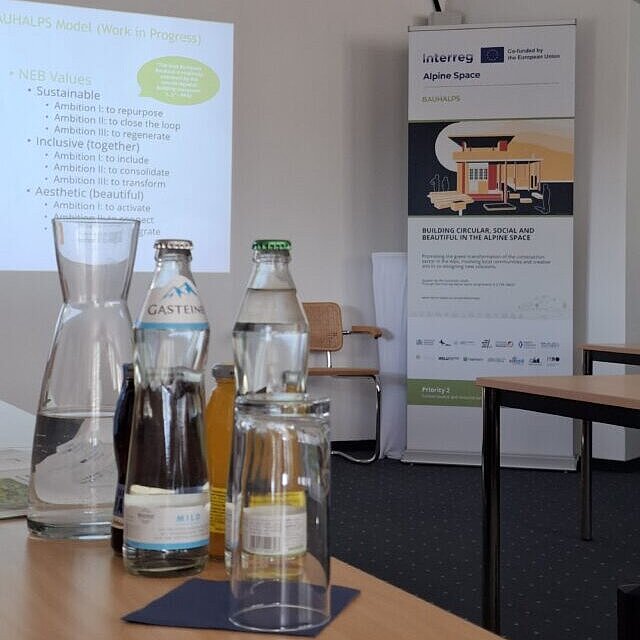 Glasflaschen auf einem Workshoptisch, dahinter eine Leinwand mit Präsentation und ein Roll-up. 