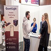 Das Bild zeigt eine Gruppe von Personen, die sich an einem Messestand oder Ausstellungsbereich unterhalten. 