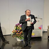 Markus Eder, Ministerialdirigent im Bayerischen Staatsministerium für Wirtschaft, Landes-entwicklung und Energie, spricht in ein Mikrofon, während hinter ihm ein Pult und ein Blu-menstrauß in einer Vase zu sehen sind.