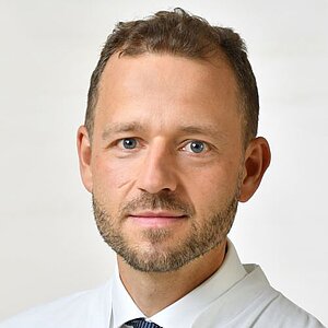 Mann mit kurzen, braunen Haaren, weißem Hemd und dunkelblauer Krawatte vor hellem Hintergrund