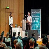 In einem Konferenz- oder Präsentationsraum sitzt ein Publikum und blickt auf eine Bühne. Auf der Bühne stehen zwei Personen: Eine spricht hinter einem Rednerpult, die andere steht seitlich neben einem Tisch mit Flaschen und Gläsern. Im Hintergrund ist ein großes Banner oder eine Leinwand mit veranstaltungsbezogenem Text zu sehen. 