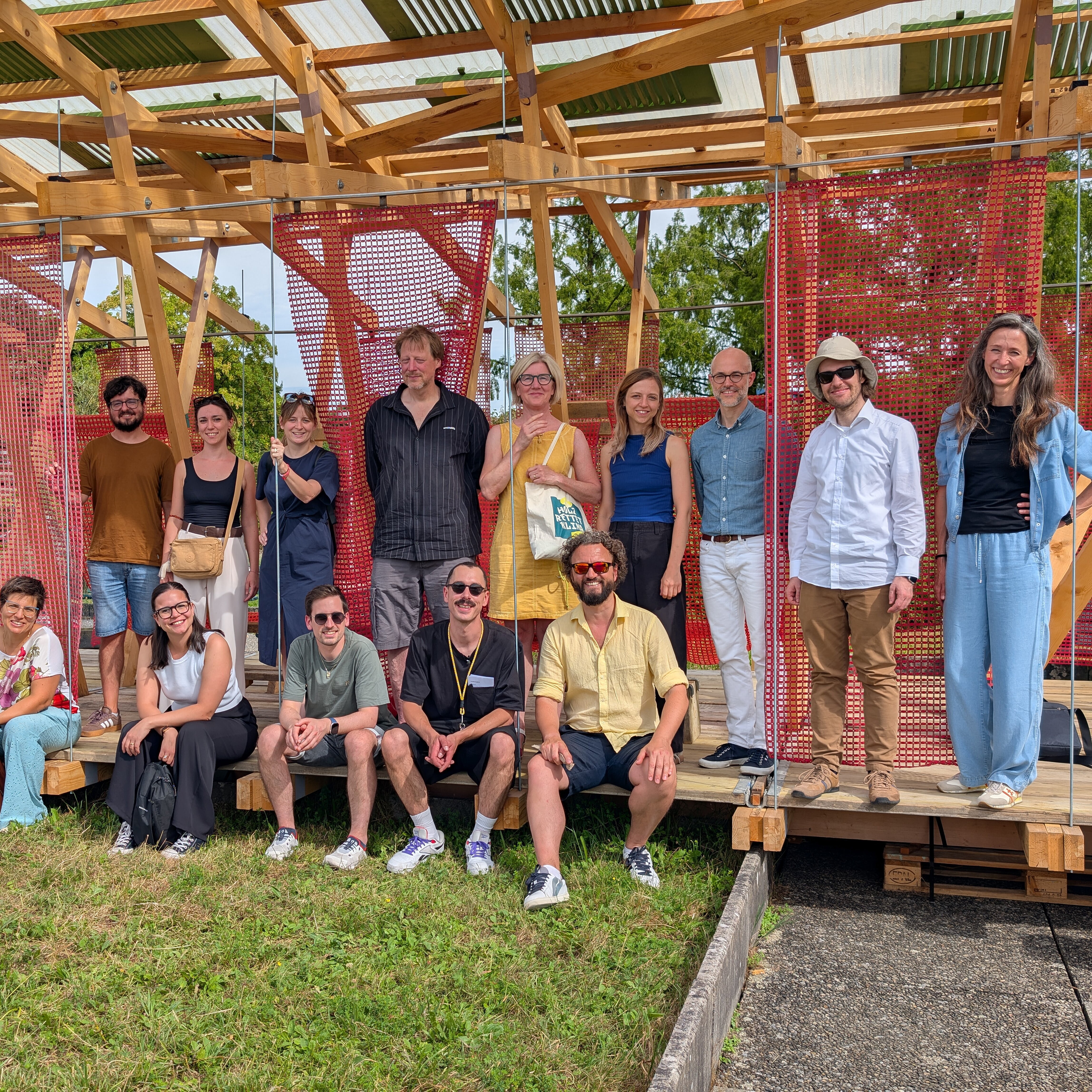 Das Bild zeigt eine Gruppe von Menschen, die vor einer offenen Holzkonstruktion mit roten Netzelementen für ein gemeinsames Foto posiert. Einige stehen, andere sitzen auf einer Holzplattform; im Hintergrund sind Bäume, Berge und ein teilweise bewölkter Himmel zu sehen.