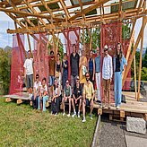 Das Bild zeigt eine Gruppe von Menschen, die vor einer offenen Holzkonstruktion mit roten Netzelementen für ein gemeinsames Foto posiert. Einige stehen, andere sitzen auf einer Holzplattform; im Hintergrund sind Bäume, Berge und ein teilweise bewölkter Himmel zu sehen.