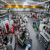 Blick in eine große Industriehalle mit mehreren Maschinen, Regalen und einem roten Industrieroboter unter einer Decke mit Rohrleitungen und Beleuchtung