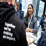 Eine Frau trägt einen Hoodie und redet mit einem Mann, der am Tisch sitzt.