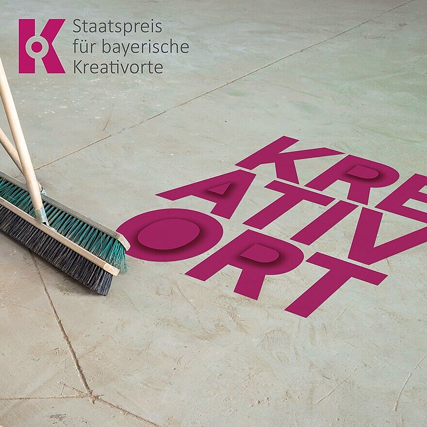 Das Bild zeigt eine glatte Betonfläche, auf der in großen, kräftigen pinken Buchstaben das Wort „KREATIVORT“ steht. Links lehnen zwei Besen an einer Wand, die Borsten berühren den Boden. Oben links ist das Logo der Auszeichnung „Staatspreis für bayerische Kreativorte“ zu sehen, bestehend aus einem stilisierten „K“ in Pink und dem Schriftzug daneben. Der Bildaufbau vermittelt den Eindruck eines vorbereiteten oder aufgeräumten Raumes, der mit Kreativität in Verbindung gebracht wird.