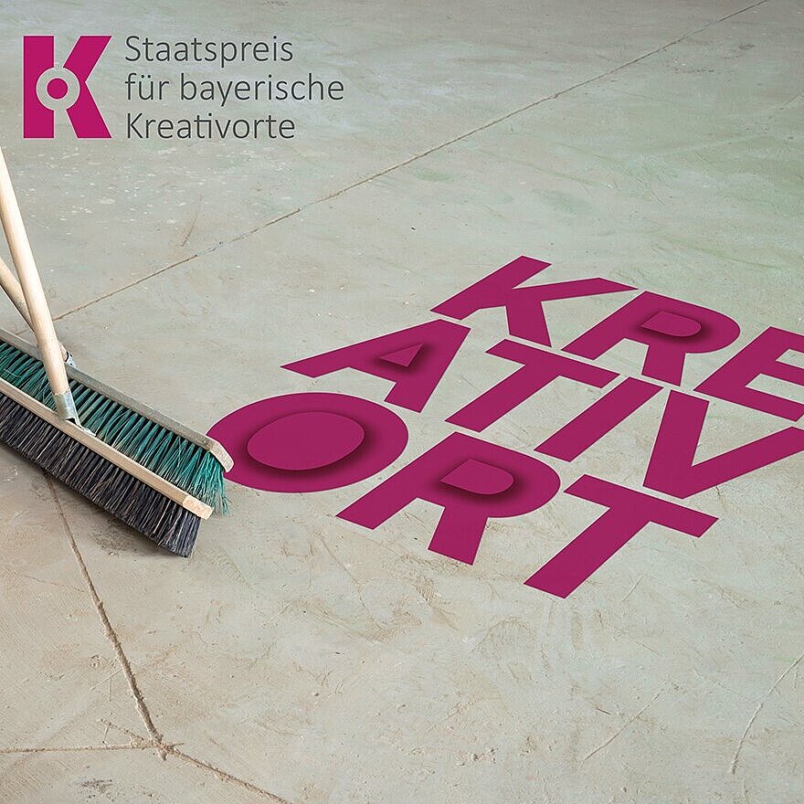 Das Bild zeigt eine glatte Betonfläche, auf der in großen, kräftigen pinken Buchstaben das Wort „KREATIVORT“ steht. Links lehnen zwei Besen an einer Wand, die Borsten berühren den Boden. Oben links ist das Logo der Auszeichnung „Staatspreis für bayerische Kreativorte“ zu sehen, bestehend aus einem stilisierten „K“ in Pink und dem Schriftzug daneben. Der Bildaufbau vermittelt den Eindruck eines vorbereiteten oder aufgeräumten Raumes, der mit Kreativität in Verbindung gebracht wird.