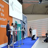 „Eine Gruppe von Personen steht auf einer Bühne mit blauem Boden und weißen Wänden. Im Hintergrund befindet sich ein großer Bildschirm mit Texten und Grafiken sowie mehrere Banner oder Poster. Eine Person spricht am Rednerpult, während andere daneben stehen – die Szene wirkt wie eine Veranstaltung oder Konferenz.“