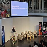 Designing PiB – Die Suche nach dem perfekten Du: Prof. Daniel Nusser, Professor und Studi-engangsleitung für Prozess- und Produktdesign, DIPLOMA Hochschule, ist zu sehen, wie er am Pult ins Mikrofon vor Publikum spricht.
