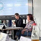 2 weibliche Personen sitzen an einem Tisch, im Hintergrund ist eine Powerpointpräsentation. 