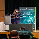 Das Bild zeigt eine Person, die an einem Rednerpult steht und vermutlich eine Rede oder Präsentation hält.