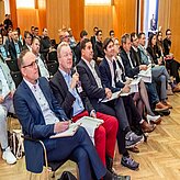 Bild zeigt eine Gruppe von Personen, die in einem Konferenz- oder Besprechungsraum sitzen. Die Anwesenden tragen formelle Kleidung wie Anzüge und Krawatten und scheinen aufmerksam zuzuhören oder sich Notizen zu machen.