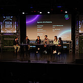 Das Bild zeigt mehrere Personen auf einer Bühne, sie sitzen alle zusammen an Tischen