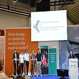 Fünf Personen stehen auf einer Bühne, jeweils hinter einem eigenen Rednerpult. Über ihnen befindet sich ein großer Bildschirm