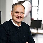 Mann mit dunklem Pullover und weißem Hemd sitzt vor einem Fenster in einem Büro oder Arbeitsraum