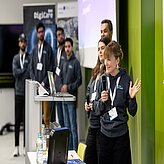 Auf dem Foto ist auf der linken Seite eine Gruppe von vier Männern zu sehen, welche vor RollUps stehen. Daneben ist eine Damit mit Mikrofon, welche einen Vortrag zu halten scheint.