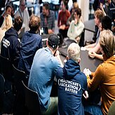 Das Bild zeigt eine Gruppe von Menschen, die an langen Tischen sitzen und an einem Healthcare Hackathon in Bayern teilnehmen. Die Teilnehmer sind in informeller Kleidung gekleidet und scheinen konzentriert an ihren Laptops zu arbeiten oder sich zu unterhalten. Auf dem Rücken eines Teilnehmers ist deutlich der Schriftzug „Healthcare Hackathon Bayern“ zu sehen.  Das Bild vermittelt den Eindruck einer aktiven und kollaborativen Arbeitsatmosphäre.