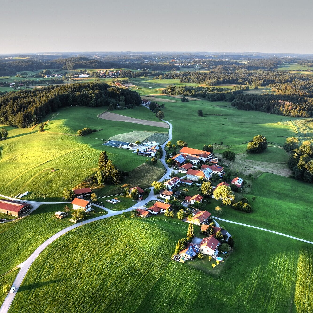 Drohnenaufnahme eines kleinen Dorfes. Drumherum befinden sich viele grüne Felder und Wälder