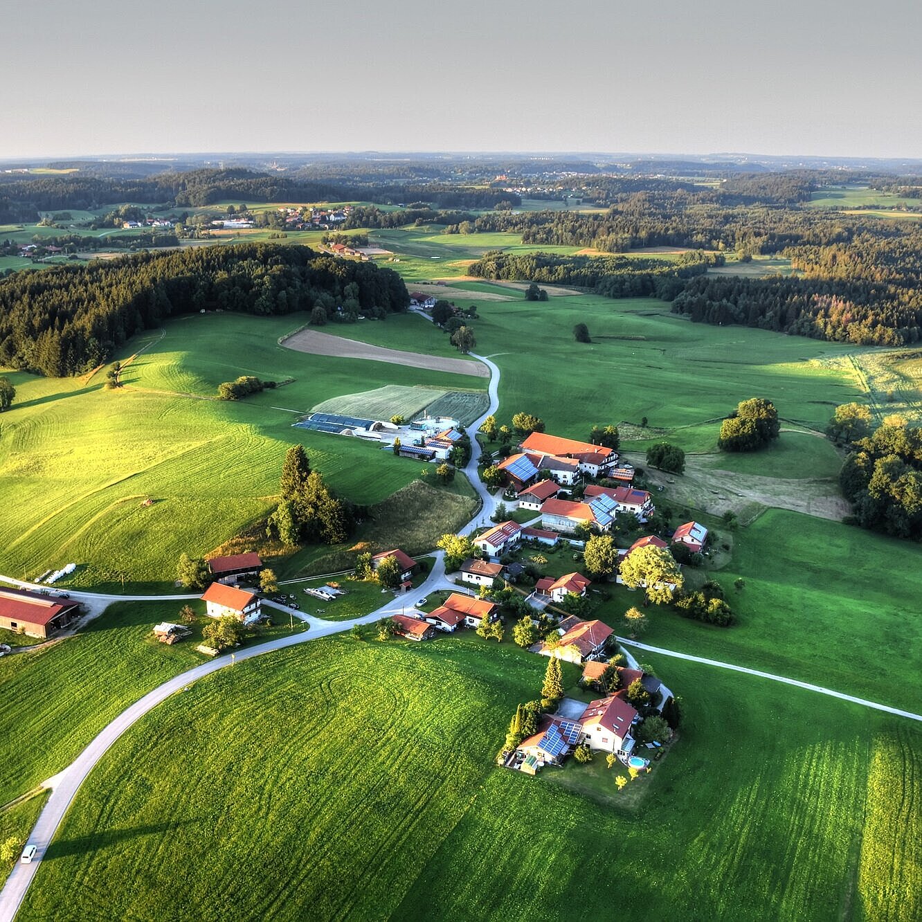 Drohnenaufnahme eines kleinen Dorfes. Drumherum befinden sich viele grüne Felder und Wälder