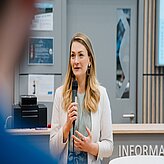 Frau mit langen blonden Haaren hält Mikrofon und spricht vor einem Informationsschalter.