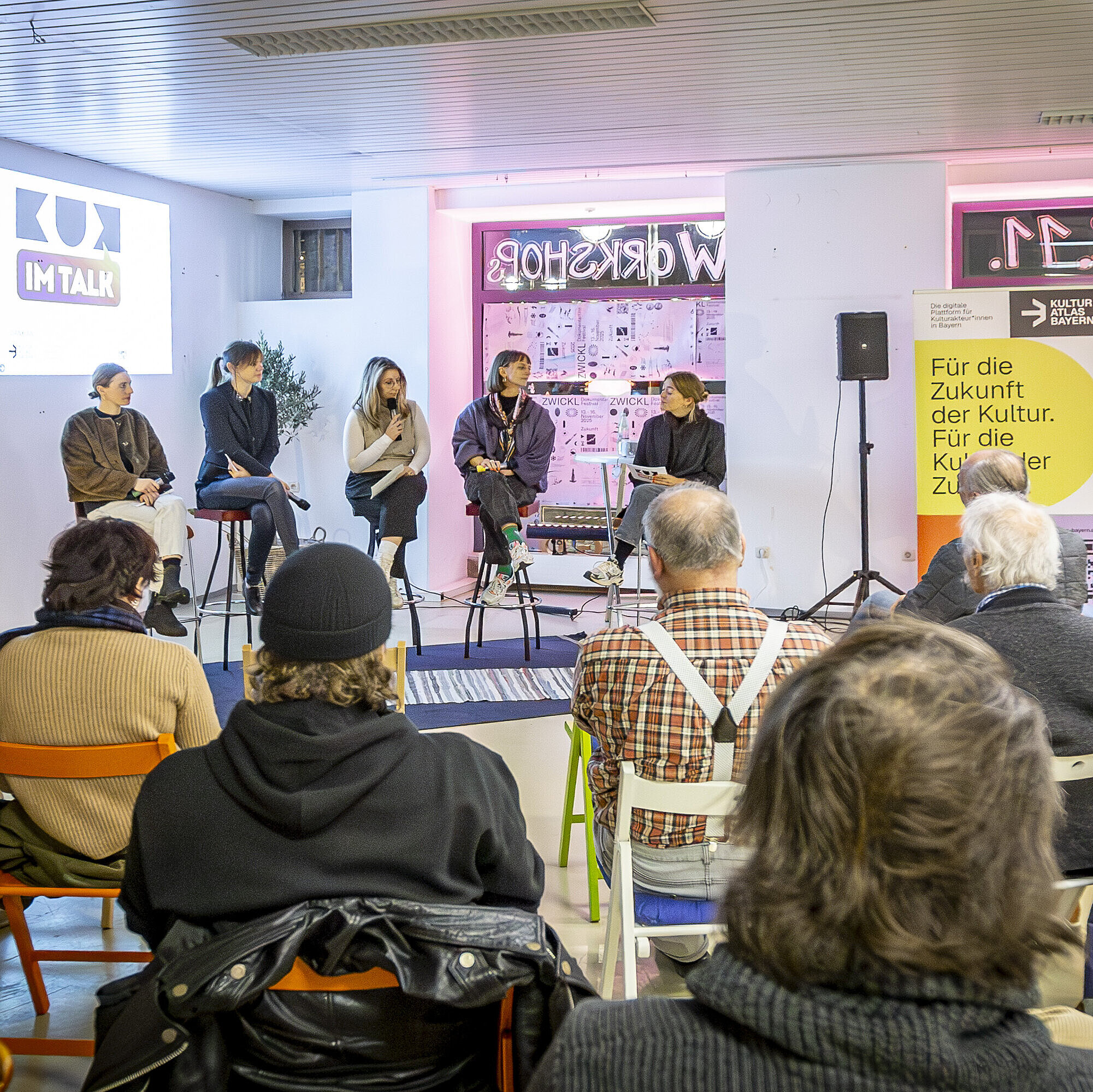 Auf dem Bild sitzt ein fünfköpfiges Panel, darunter Charlotte Stegmayer, Projektmanagerin im Innovationsnetzwerk Kreativwirtschaft der Bayern Innovativ GmbH, auf hohen Hockern vor einem Publikum. Hinter dem Panel ist eine Projektion mit dem Titel ‚KUK im Talk‘ zu sehen. Rechts steht ein gelbes Banner mit dem Schriftzug ‚Für die Zukunft der Kultur‘