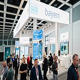 Messestand mit dem Schriftzug »bayern« auf einem weißen Banner und mehreren Bildschirmen in einer Messehalle mit mehreren Personen