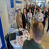 Mehrere Personen stehen an einem runden Stehtisch mit Informationsmaterial und Mikrofon in einem hellen Raum mit Plakaten an der Wand