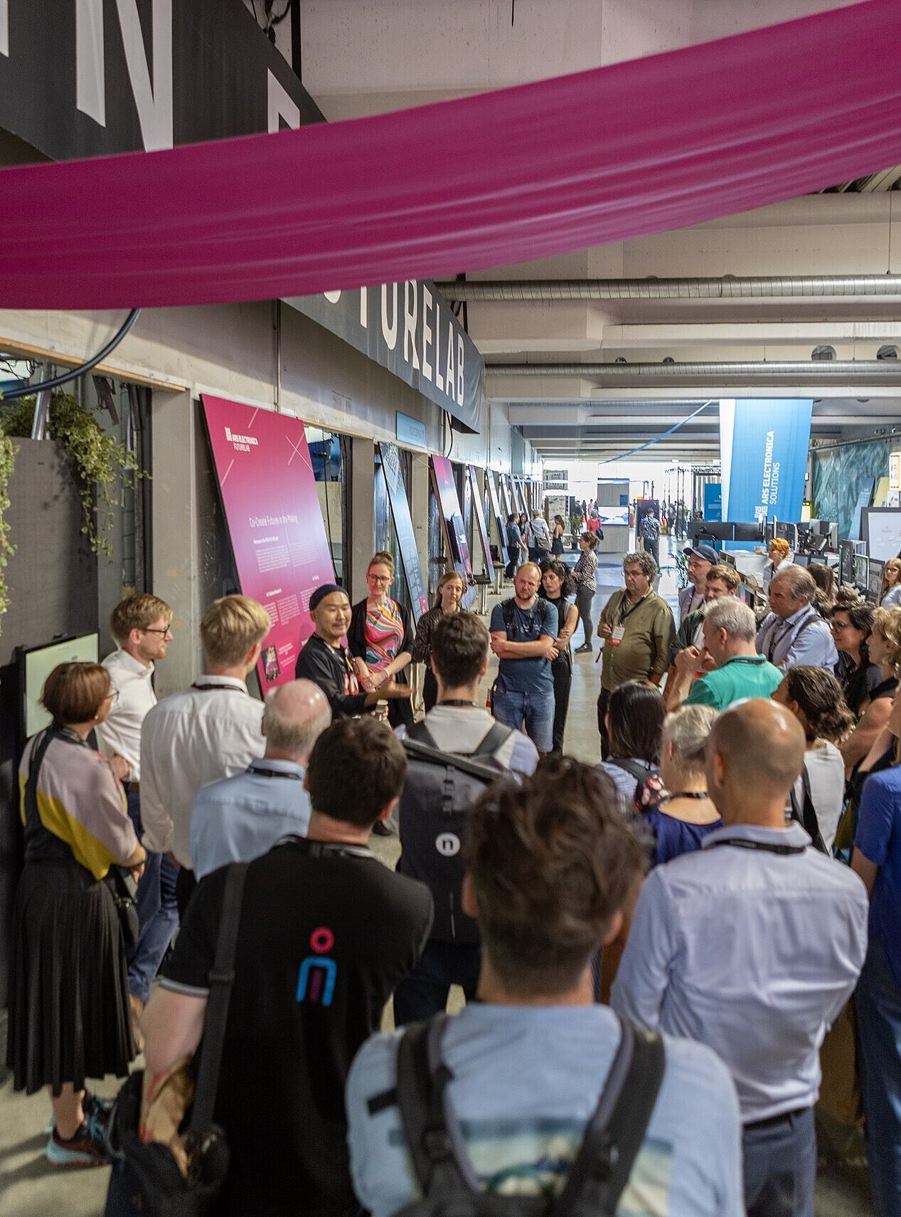Eine große Gruppe von Menschen steht in einer Halle und hört einer Person zu, die vorne etwas erklärt. An der Wand hängen mehrere bunte Infotafeln. Über den Köpfen hängt ein breites pinkes Tuch. Im Hintergrund sieht man weitere Stände und Banner.