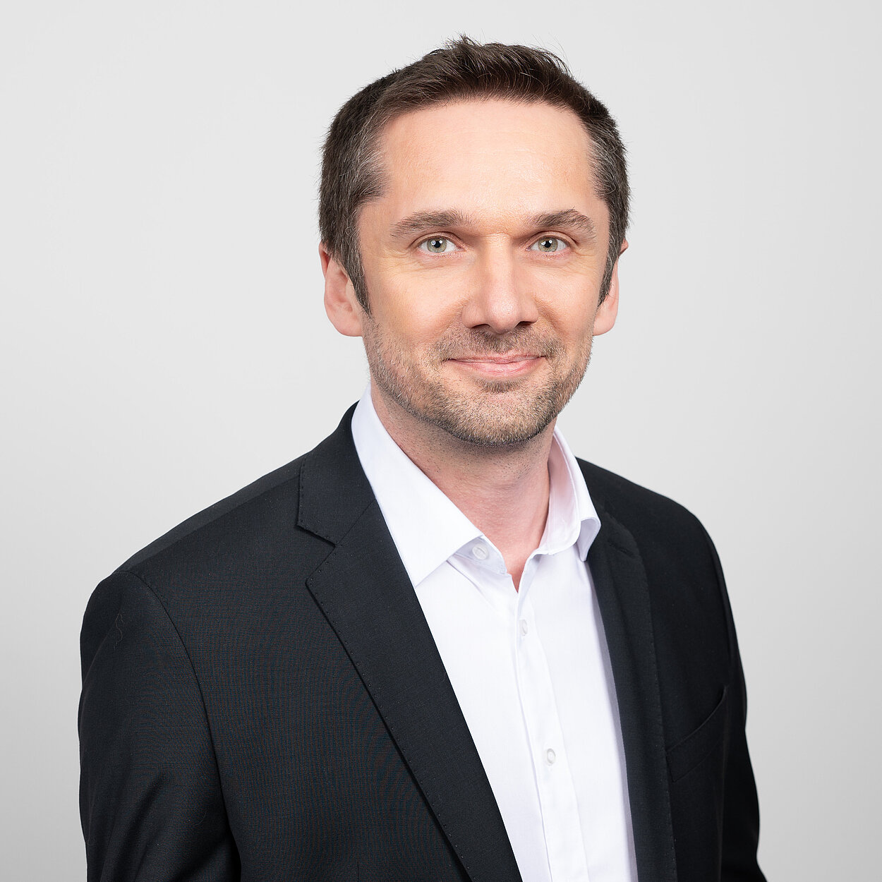 Ein dunkelhaariger Mann in einem schwarzen Anzug mit weißem Oberhemd ist vor einem hellgrauen Hintergrund abgelichtet.